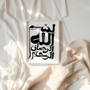 A framed black and white art print featuring Arabic calligraphy. The artwork is displayed in a white frame against a backdrop of soft, draped fabric. The calligraphy is bold and stylized, creating a striking visual contrast.