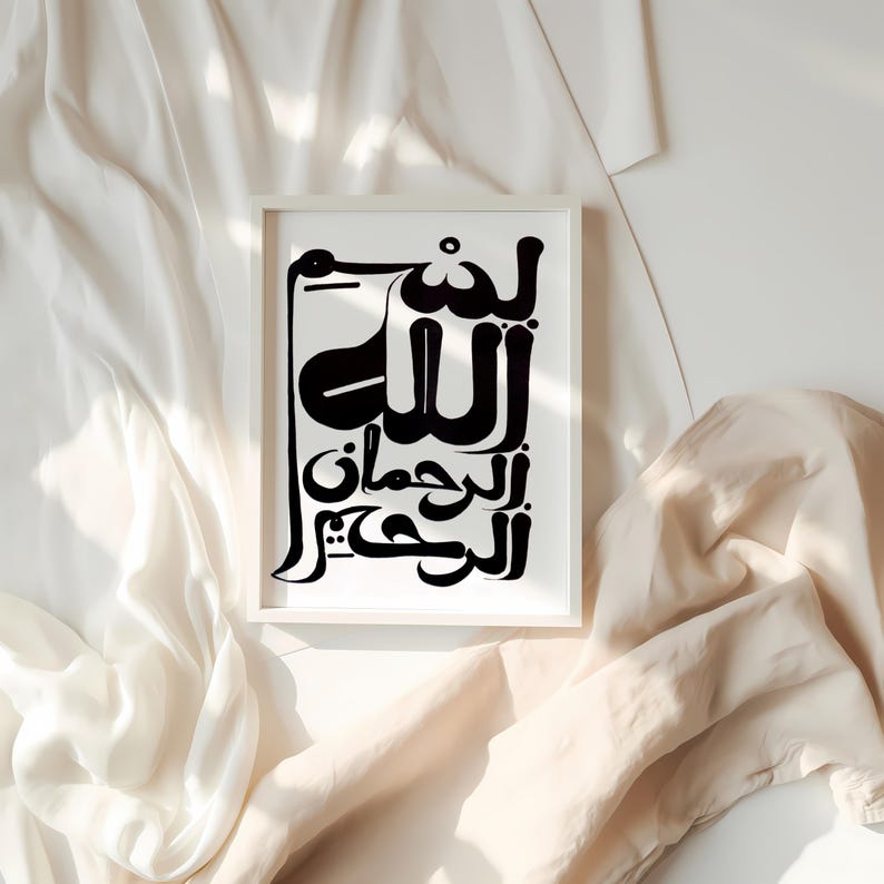A framed black and white art print featuring Arabic calligraphy. The artwork is displayed in a white frame against a backdrop of soft, draped fabric. The calligraphy is bold and stylized, creating a striking visual contrast.