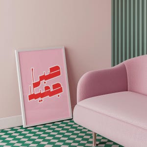 May include: A framed pink art print with red Arabic calligraphy leans against a wall. The print is in a white frame and is set against a pink wall. A pink armchair and green and white checkered rug are also visible.