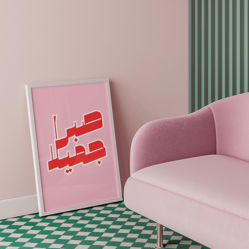 May include: A framed pink art print with red Arabic calligraphy leans against a wall. The print is in a white frame and is set against a pink wall. A pink armchair and green and white checkered rug are also visible.