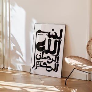 Black and white framed art print featuring Arabic calligraphy. The artwork displays elegant script on a white background, set within a sleek black frame. The print is leaning against a white wall, with a wooden floor in the foreground.