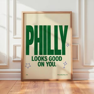 Könnte beinhalten: Ein rechteckiger Wandkunstdruck mit cremefarbenem Hintergrund und dunkelgrünem Text, der PHILLY LOOKS GOOD ON YOU lautet. Das Design enthält kleine blaue Sternsymbole, die Zahl 215 unten links und PHILADELPHIA, PA unten rechts.