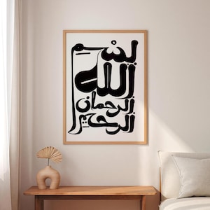A framed art print featuring black Arabic calligraphy on a white background. The artwork is in a wooden frame and hangs on a white wall. A small, beige vase with a decorative fan sits on a wooden table below the artwork.
