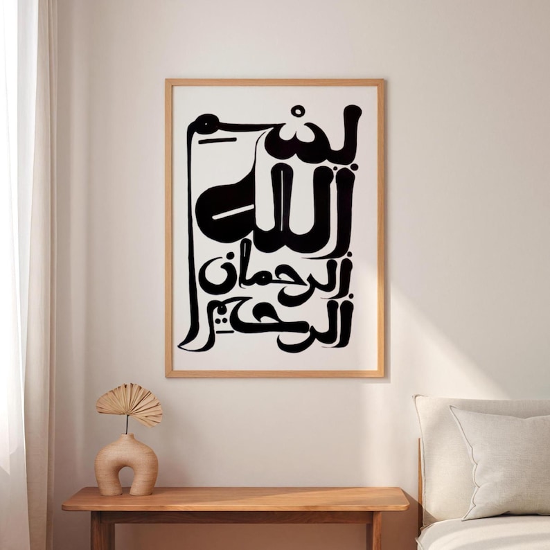 A framed art print featuring black Arabic calligraphy on a white background. The artwork is in a wooden frame and hangs on a white wall. A small, beige vase with a decorative fan sits on a wooden table below the artwork.