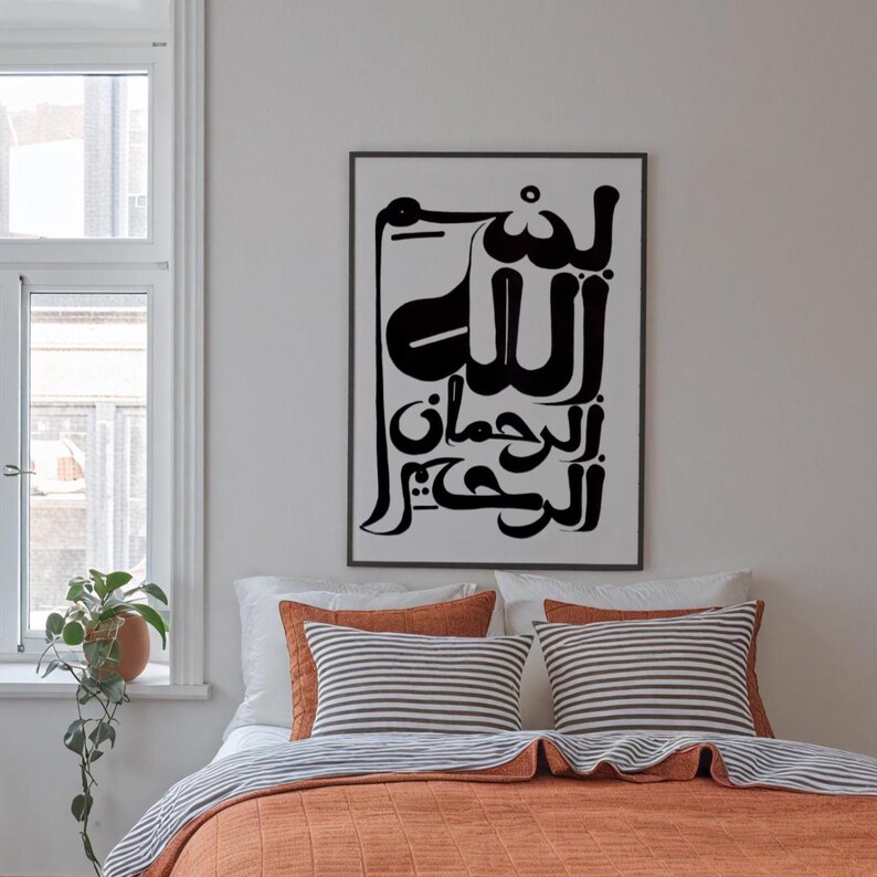 A framed black and white Arabic calligraphy art print hangs above a bed with orange bedding and striped pillows. The artwork features bold, stylized script. The bedroom has a window with a potted plant.
