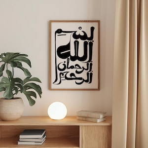 A framed art print featuring black Arabic calligraphy on a white background. The artwork is in a wooden frame and hangs on a white wall. A potted plant, a glowing orb lamp, and books sit on a wooden shelf below.