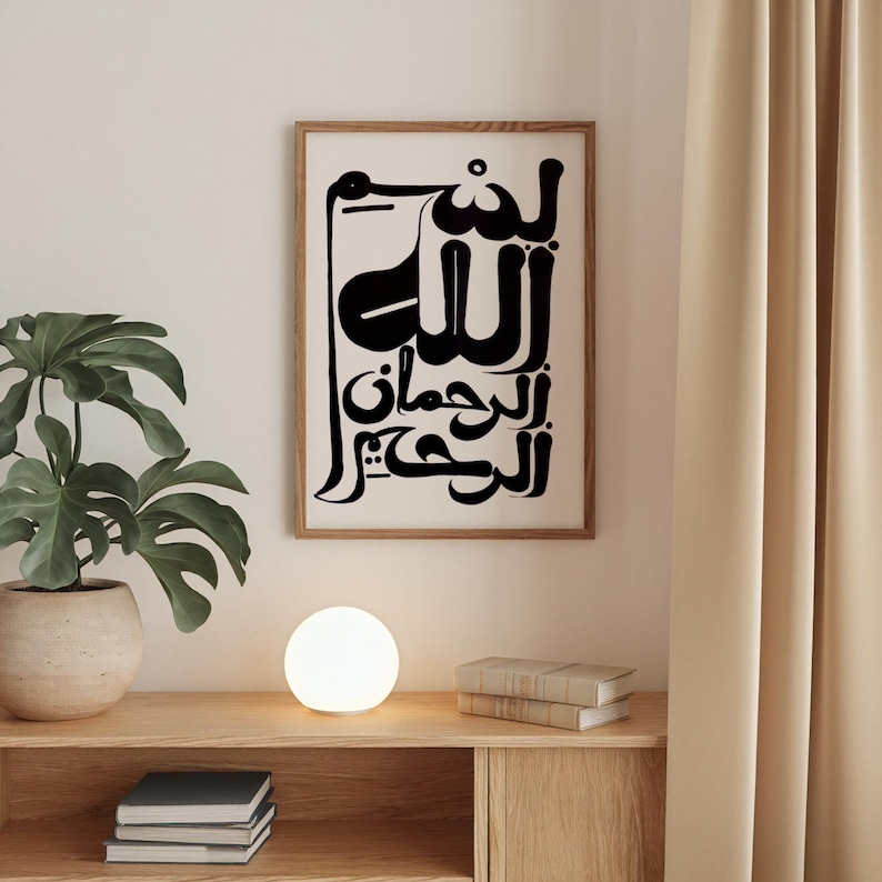 A framed art print featuring black Arabic calligraphy on a white background. The artwork is in a wooden frame and hangs on a white wall. A potted plant, a glowing orb lamp, and books sit on a wooden shelf below.