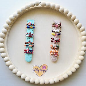May include: A set of colorful beaded rings with football and letter charms. The rings are displayed on two wooden stands with a heart-shaped sticker that says "Lil' Shay Designs".