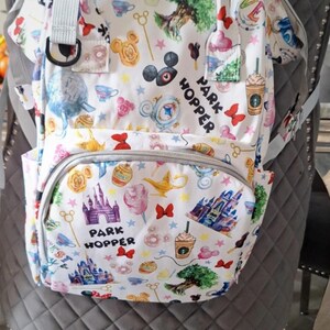May include: White backpack with a colourful pattern featuring cartoon characters, castles, and the text "PARK HOPPER." The backpack has a front pocket, side pockets, and a top handle. The bag is sitting on a grey chair.