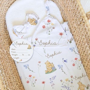 May include: A white baby swaddle blanket, hat, and head support with a Winnie the Pooh and floral pattern, personalized with the name "Sophia." The set is nestled in a woven basket, with a matching tag for recording birth details.