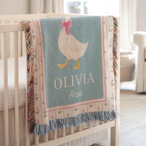 May include: A baby blanket with a light blue background and a white goose design wearing a pink bow. The blanket has the name "OLIVIA Rae" in white letters. The blanket is draped over a white crib.