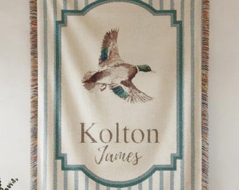 May include: A beige and blue woven throw blanket with a duck illustration. The blanket features vertical blue stripes and a teal border. The name "Kolton James" is printed on the blanket.