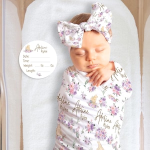 May include: A newborn baby wrapped in a white floral swaddle blanket and matching headband. The blanket and headband feature a pattern of purple flowers and cartoon characters. A circular tag with the name "Adeline Rylee" is visible.