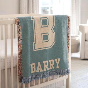 May include: A teal baby blanket with the letter "B" and the name "BARRY" in a cream-colored, collegiate-style font. The blanket has a fringed edge and is draped over a white crib. The nursery has a neutral color palette.