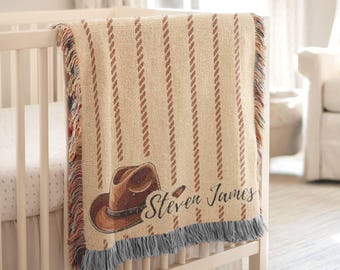 May include: A beige and brown baby blanket featuring a cowboy hat design and the name "Steven James". The blanket has vertical brown stripes and a fringed edge. It is draped over a white cot.