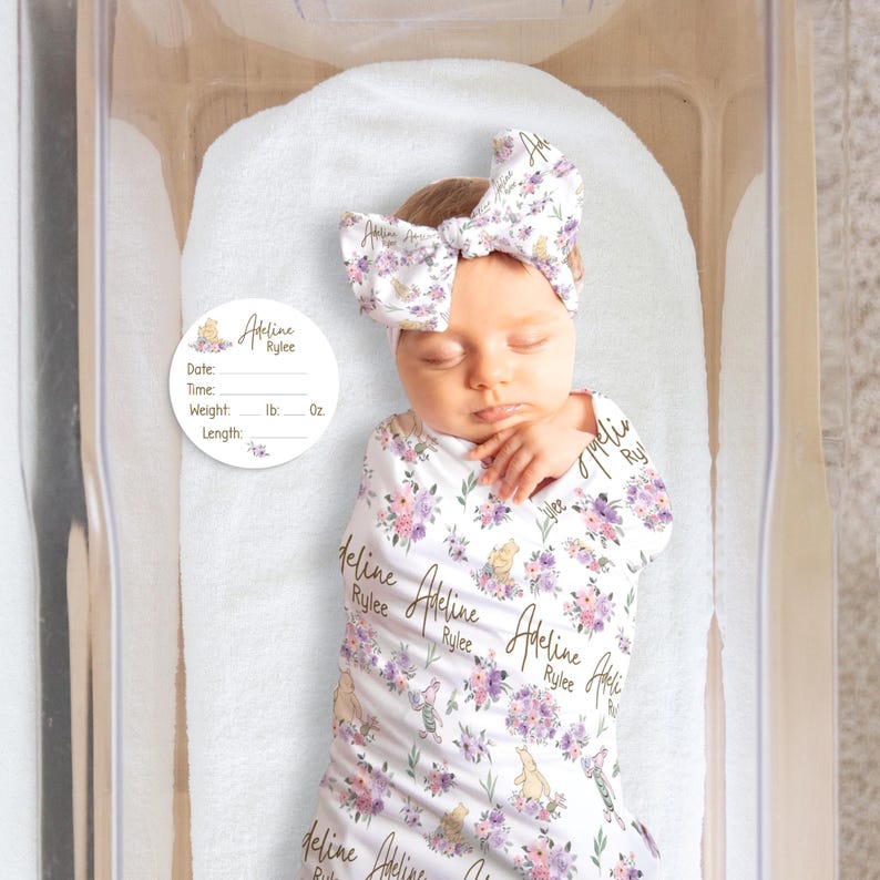 May include: A newborn baby wrapped in a white floral swaddle blanket and matching headband. The blanket and headband feature a pattern of purple flowers and cartoon characters. A circular tag with the name "Adeline Rylee" is visible.