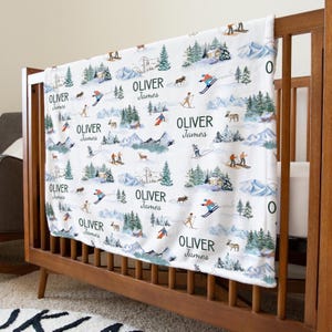 May include: White baby blanket with a winter ski scene design. The blanket features skiers, trees, mountains, and the text "OLIVER James" repeated across the surface. The blanket is draped over a wooden crib.