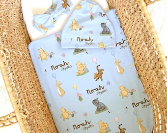 May include: A light blue baby swaddle blanket, knotted hat, and bow headband set featuring a repeating pattern of Winnie the Pooh characters, balloons, and the printed name Noah Jayden in a dark brown script font throughout the fabric.
