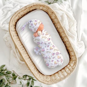 May include: A newborn baby swaddled in a white floral print receiving blanket and matching hat, nestled in a wicker bassinet. The floral pattern includes purple and yellow flowers. The baby is holding a small stuffed animal.