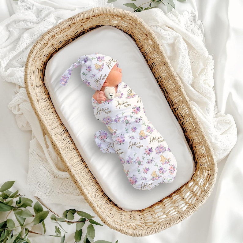 May include: A newborn baby swaddled in a white floral print receiving blanket and matching hat, nestled in a wicker bassinet. The floral pattern includes purple and yellow flowers. The baby is holding a small stuffed animal.