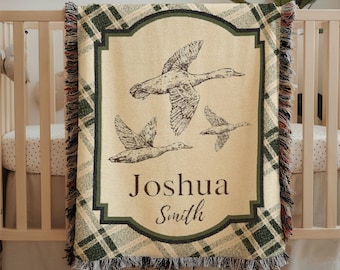 May include: A woven baby blanket with a cream-colored center featuring three flying ducks. The center is framed by a green border and a plaid pattern. The name "Joshua Smith" is printed below the ducks. The blanket has a fringed edge.