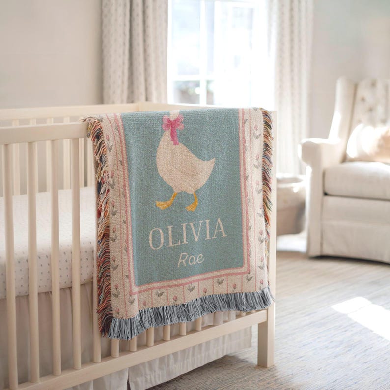 May include: A baby blanket with a light blue background and a white goose design wearing a pink bow. The blanket has the name "OLIVIA Rae" in white letters. The blanket is draped over a white crib.