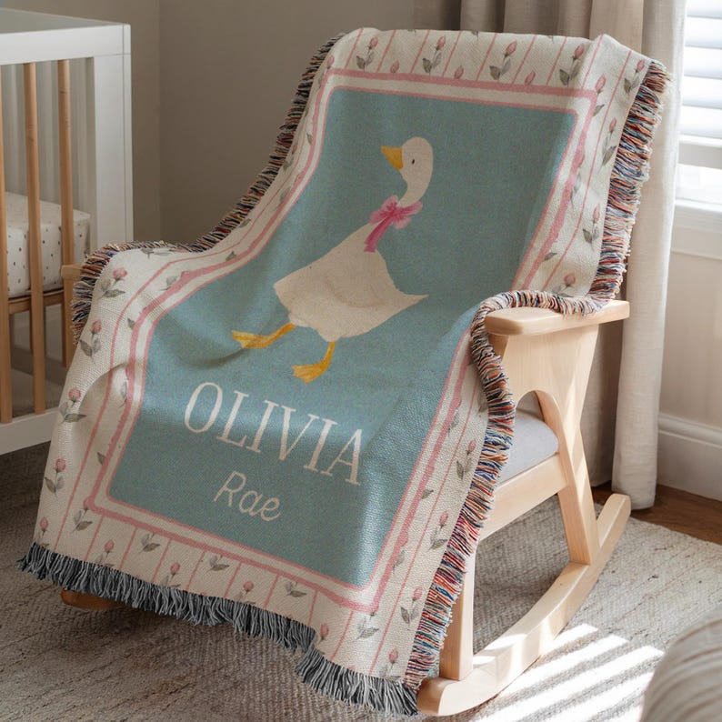 May include: A light blue and white woven baby blanket with a goose illustration and the name "OLIVIA Rae". The blanket has a pink floral border and fringe trim. The blanket is draped over a wooden rocking chair.
