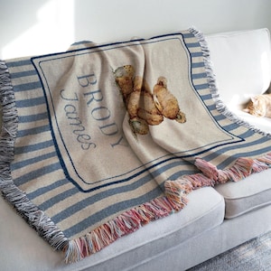 May include: A beige throw blanket with a teddy bear illustration and the name "BRODY James" in blue script. The blanket has blue and white stripes and a fringed edge. The blanket is draped over a white sofa.