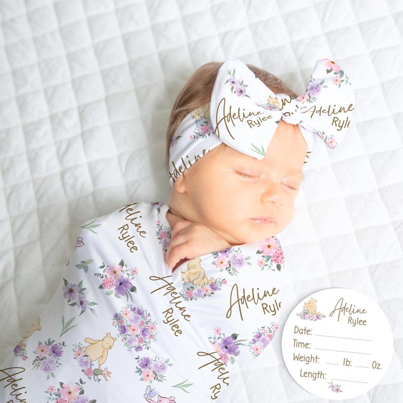 May include: A newborn baby wrapped in a white floral swaddle blanket and matching headband. The blanket and headband feature a purple and pink floral pattern with a Winnie the Pooh design and the name "Adeline Rylee". A round name announcement card is next to the baby.