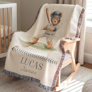 May include: A cream-coloured baseball-themed throw blanket featuring a teddy bear batter illustration. The blanket has a baseball stitch design and the name "LUCAS James" in blue. The edges are trimmed with a multi-coloured fringe.