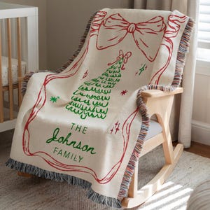 May include: Cream-colored woven throw blanket with a red Christmas tree and bow design. The blanket has a fringed edge and the words "THE Johnson FAMILY" in green. The blanket is draped over a wooden rocking chair.
