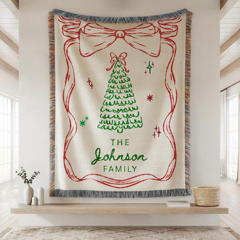 May include: A cream-colored woven throw blanket with a red bow and ribbon design framing a green Christmas tree. The words "THE JOHNSON FAMILY" are written in green script. The blanket has a gray fringe.