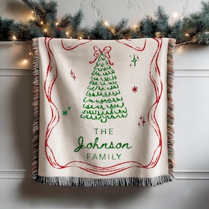 May include: Cream-colored woven throw blanket with a red border and fringe. A green Christmas tree design is centered, with the text "THE JOHNSON FAMILY" in green. Festive holiday decor.