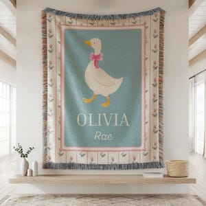 May include: A woven throw blanket featuring a white goose with a pink bow on a light blue background. The blanket has a pink and floral border, with the name "OLIVIA Rae" printed below the goose. The blanket has a fringed edge.