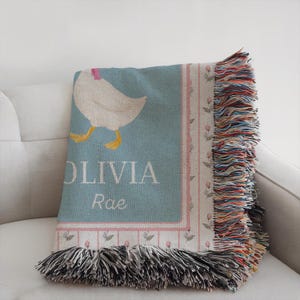 May include: A light blue throw blanket with a white goose illustration and the name "OLIVIA Rae" in white text. The blanket has a pink floral border and colorful fringe.