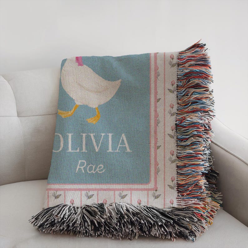 May include: A light blue throw blanket with a white goose illustration and the name "OLIVIA Rae" in white text. The blanket has a pink floral border and colorful fringe.