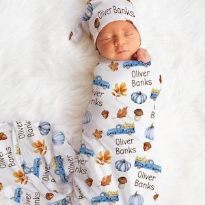 May include: A newborn baby wrapped in a white swaddle and matching hat, both adorned with a fall-themed pattern. The pattern includes blue trucks, pumpkins, leaves, and the name "Oliver Banks".