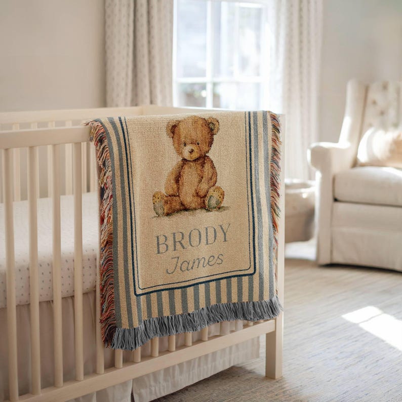 May include: A beige baby blanket with a brown teddy bear illustration and the name "BRODY James" in blue lettering. The blanket has blue and white striped borders and a fringed edge. The blanket is draped over a white crib.