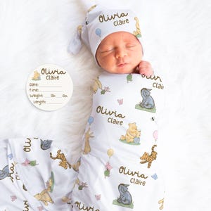 May include: A white baby swaddle blanket and matching hat featuring Winnie the Pooh characters. The blanket and hat are printed with the name "Olivia Claire". A circular tag is also present with the same name and space for date, time, weight, and length.