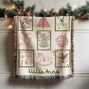 May include: A cream-colored Christmas throw blanket with red and green illustrations of festive stamps. The blanket features a Christmas tree, bow, rocking horse, and other holiday designs. The name "Lillie Anne" is printed at the bottom.