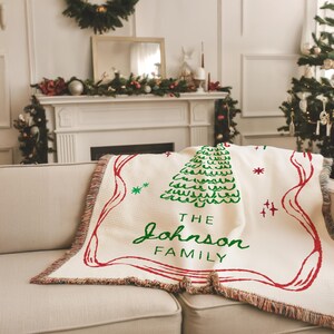 May include: Cream-colored woven throw blanket with a green Christmas tree design and red accents. The blanket features the text "THE Johnson FAMILY" in green script. The blanket is draped over a beige sofa.