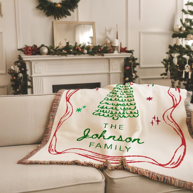 May include: Cream-colored woven throw blanket with a green Christmas tree design and red accents. The blanket features the text "THE Johnson FAMILY" in green script. The blanket is draped over a beige sofa.