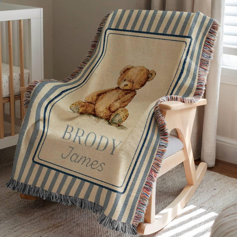 May include: A beige and blue striped baby blanket with a teddy bear illustration and the name "BRODY James". The blanket has a fringed edge and is draped over a wooden rocking chair.