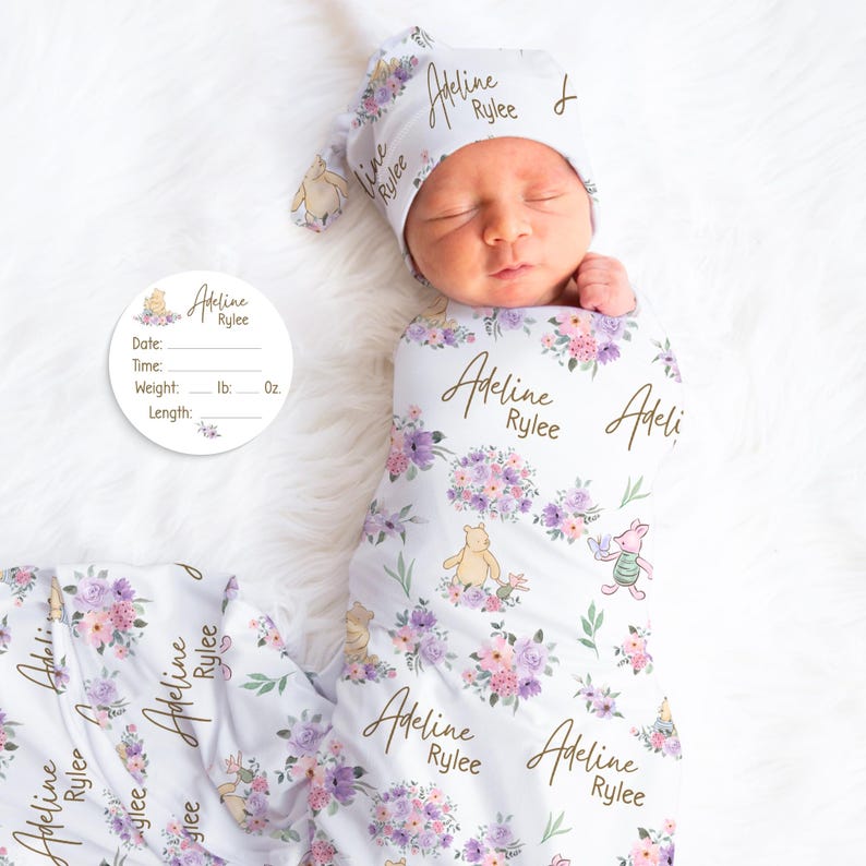 May include: A newborn baby wrapped in a white swaddle blanket and matching hat, both adorned with floral patterns, Winnie the Pooh characters, and the name "Adeline Rylee." A circular card with the same name and space for birth details is also present.