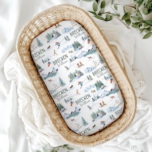 May include: A white fitted crib sheet with a mountain and skier pattern. The sheet is inside a woven basket. The pattern includes skiers, trees, deer, and the text "Brecken William" in green.