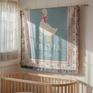 May include: A light blue baby blanket with a white goose wearing a pink bow. The blanket has the name "OLIVIA Rae" in white text. The border has a pink floral design and fringe.