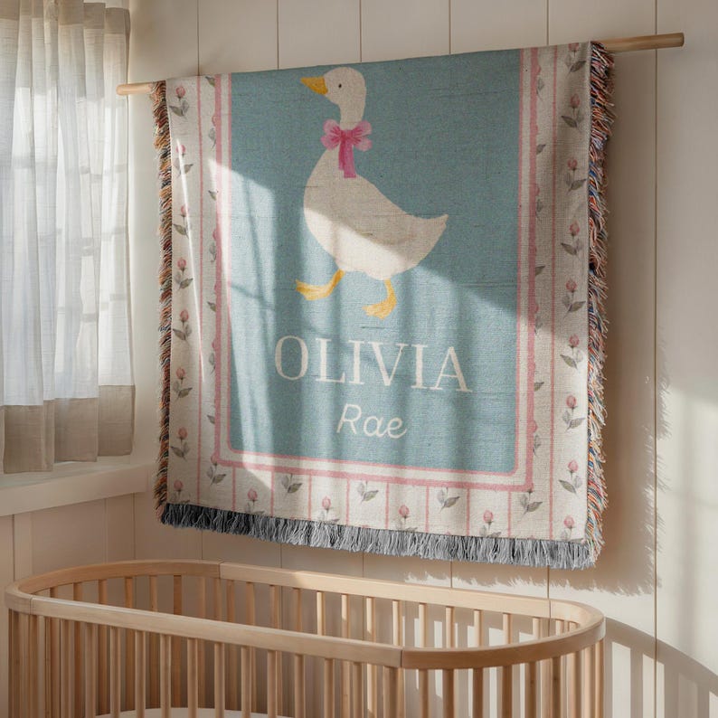 May include: A light blue baby blanket with a white goose wearing a pink bow. The blanket has the name "OLIVIA Rae" in white text. The border has a pink floral design and fringe.
