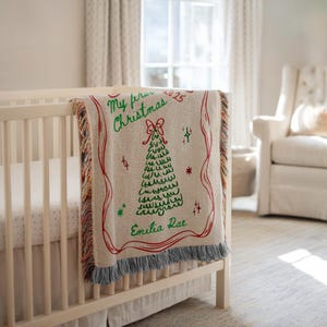 May include: A beige Christmas blanket with a red border and a green Christmas tree design. The blanket has the text "My first Christmas 2025" and the name "Emilia Rae" in green. The blanket is draped over a white crib.