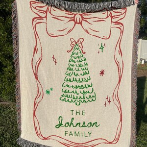 May include: Cream-colored woven throw blanket with a red bow and Christmas tree design. The blanket has a fringed edge and the words "THE Johnson FAMILY" in green script.
