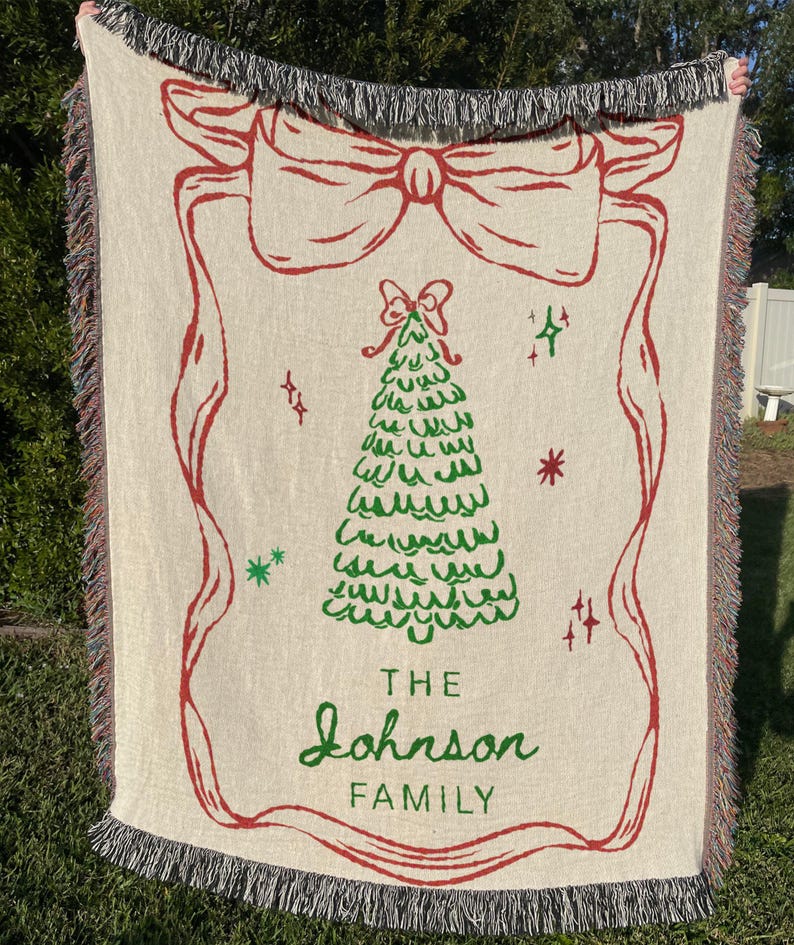 May include: Cream-colored woven throw blanket with a red bow and Christmas tree design. The blanket has a fringed edge and the words "THE Johnson FAMILY" in green script.